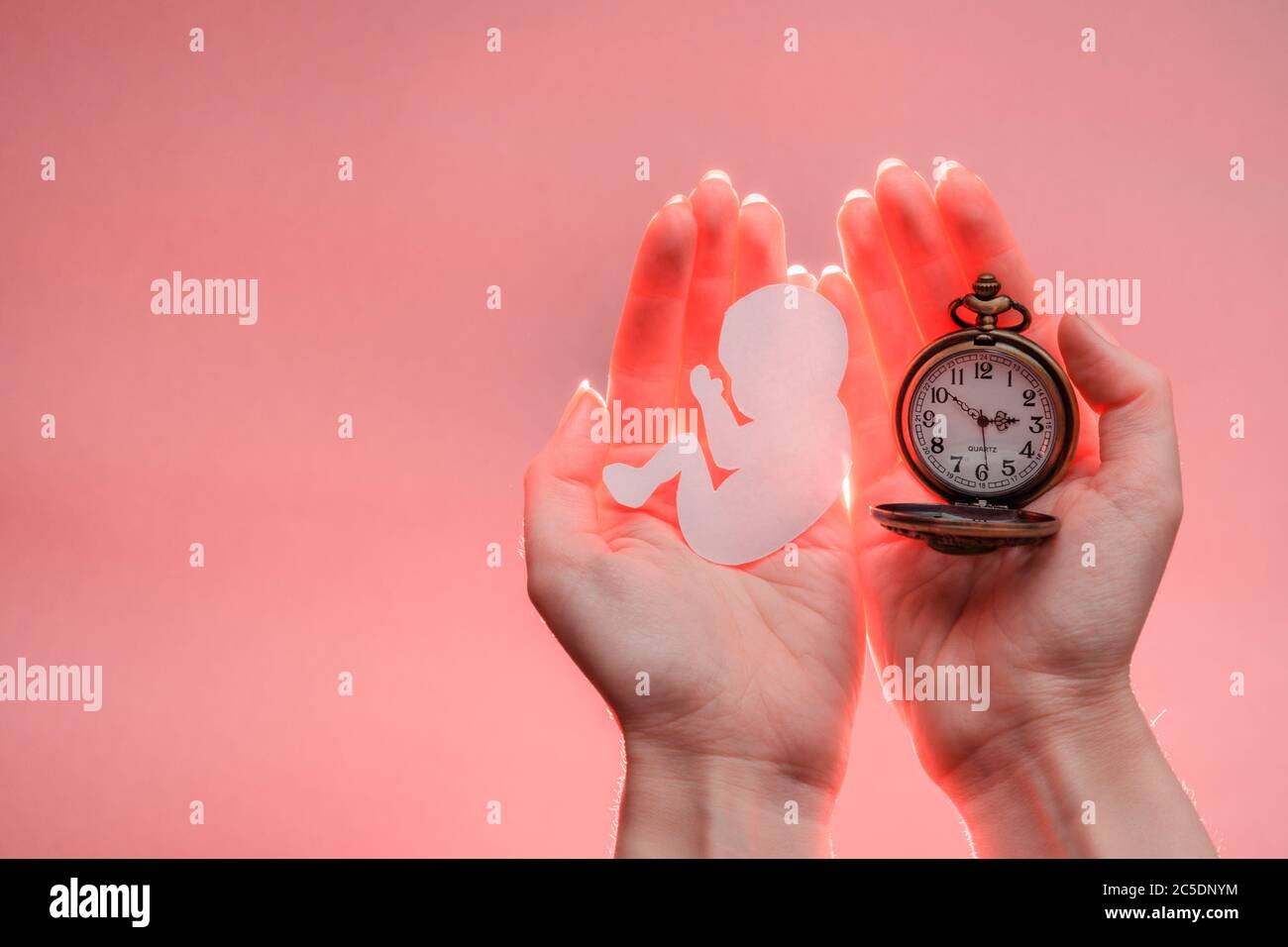 Papier silhouette d'embryon et horloge dans les mains de femme avec lumière. Arrière-plan corail clair avec espace de copie. Les mains sont sur le côté droit. Mise au point douce. Banque D'Images
