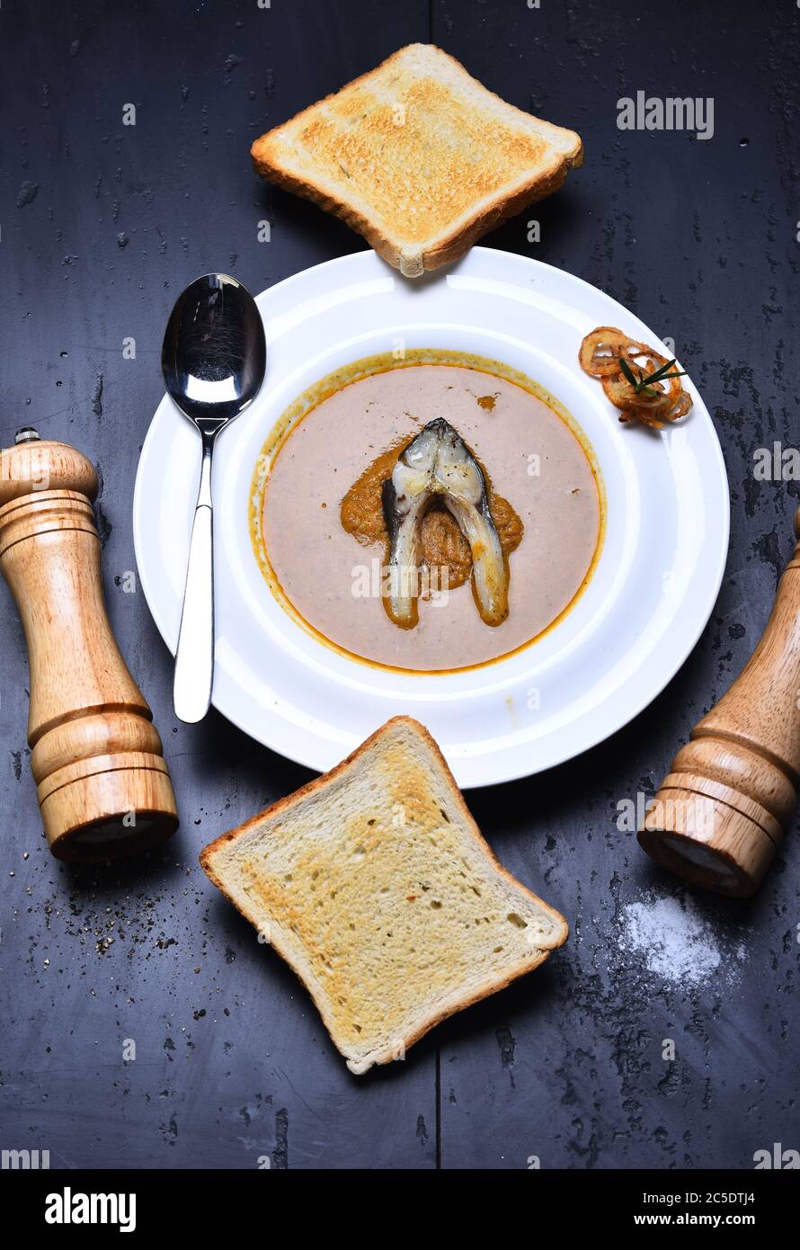 Soupe à la crème ou purée brune sur fond gris. Griller les morceaux de pain, la boîte à poivre, la cave à sel et la cuillère sur le plat. Concept de cuisine française. Soupe de poisson avec morceau de poisson et rondelles d'oignon sur plaque blanche Banque D'Images
