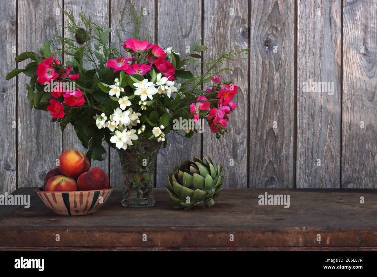 Un vase de fleurs d'été à côté d'un bol en céramique fait main rempli de pêches et d'un artichaut Banque D'Images