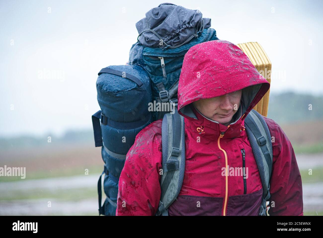 Guy randonneur touristique avec sac à dos et tapis de voyage, dans une veste rouge se tient dans la pluie Banque D'Images