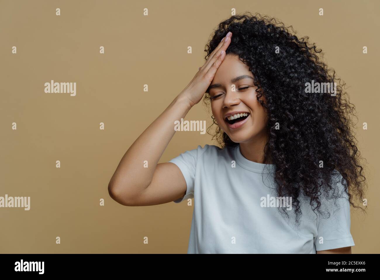 Plan horizontal de bonne positive Afro-américaine femme a plaisir, garde la main sur le front, rigole heureusement, ferme les yeux, a des cheveux bouclés, porte casua Banque D'Images