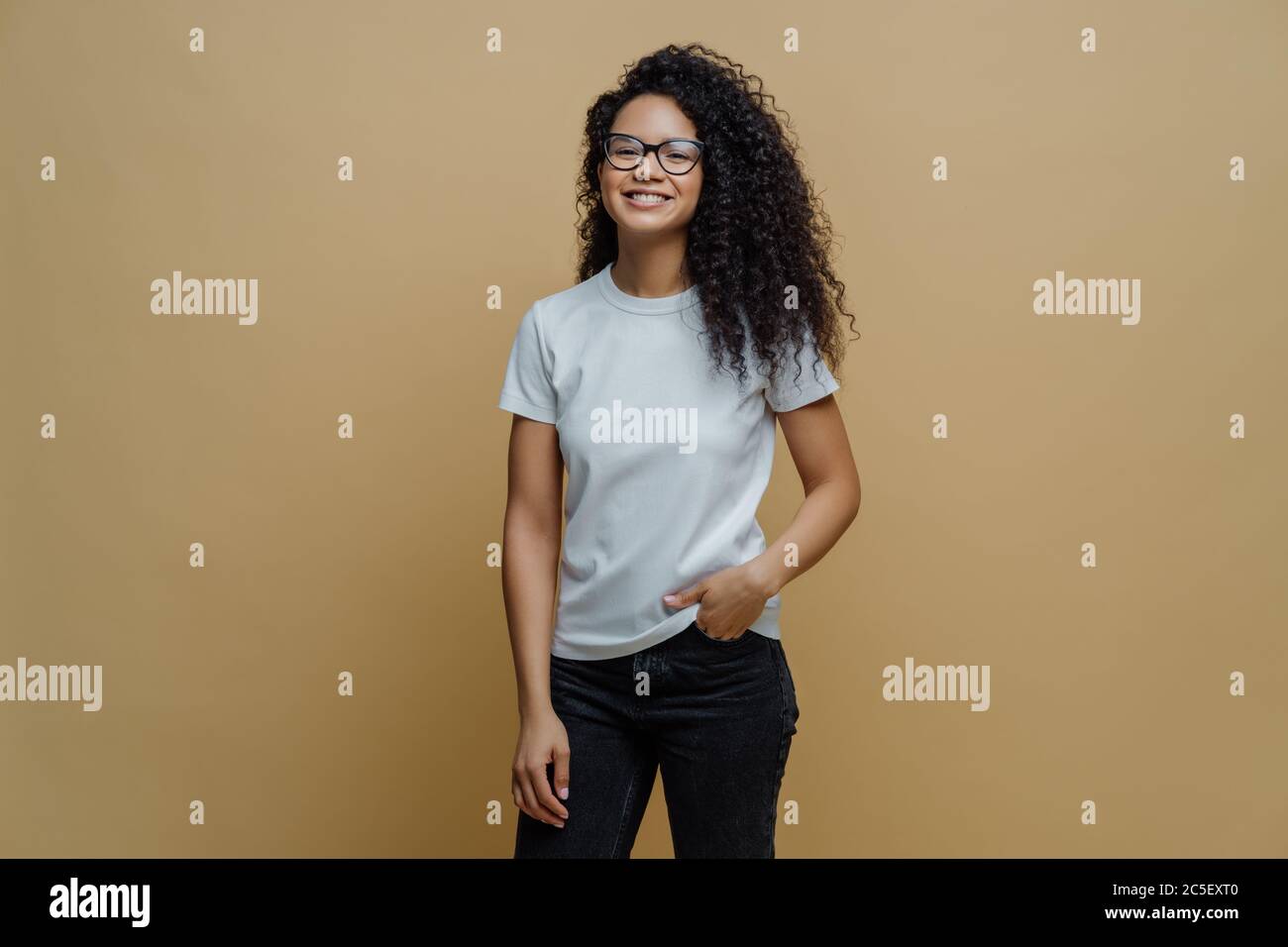 Une étudiante féminine positive se sent libre après avoir passé l'examen, sourit largement, tient la main dans la poche des jeans, porte des lunettes transparentes, se pose contre Banque D'Images