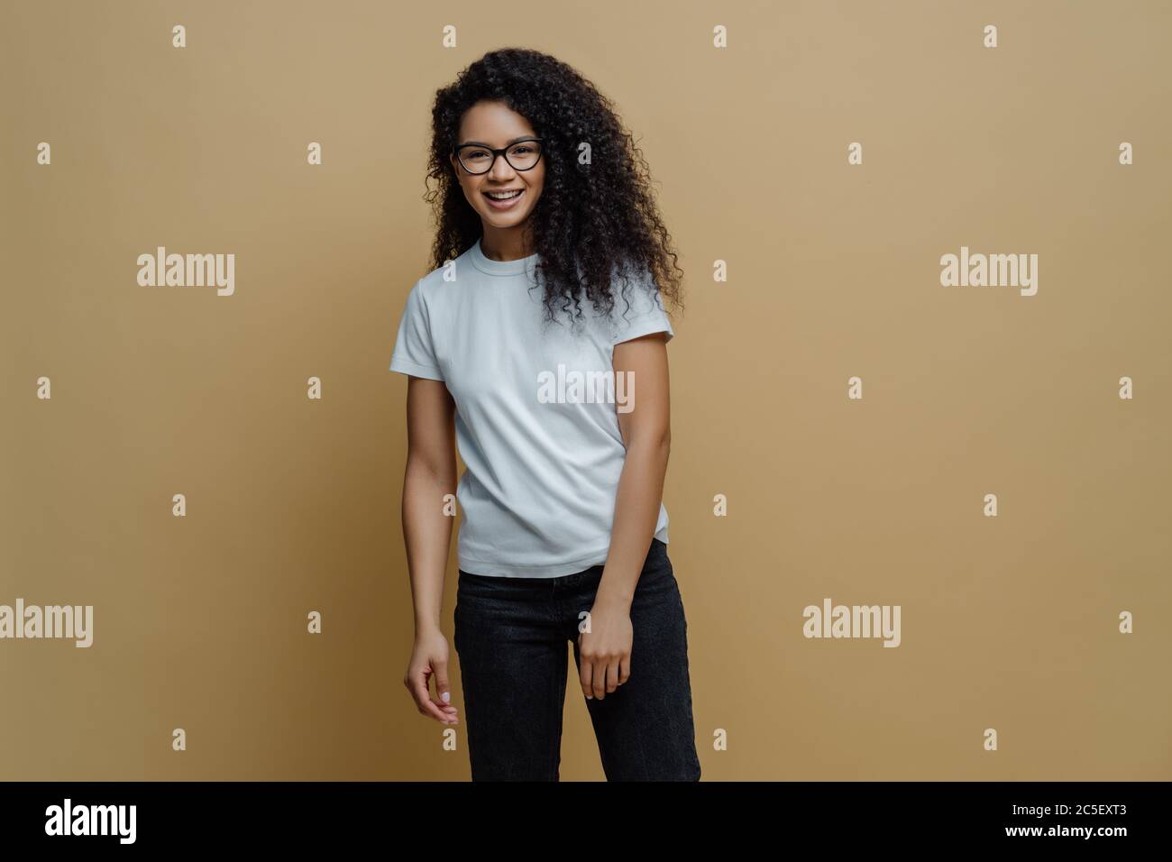 Jeune femme à la peau sombre et souriante, avec une expression du visage, porte des lunettes optiques, un t-shirt blanc et un Jean, pose sans souci sur fond beige. P Banque D'Images
