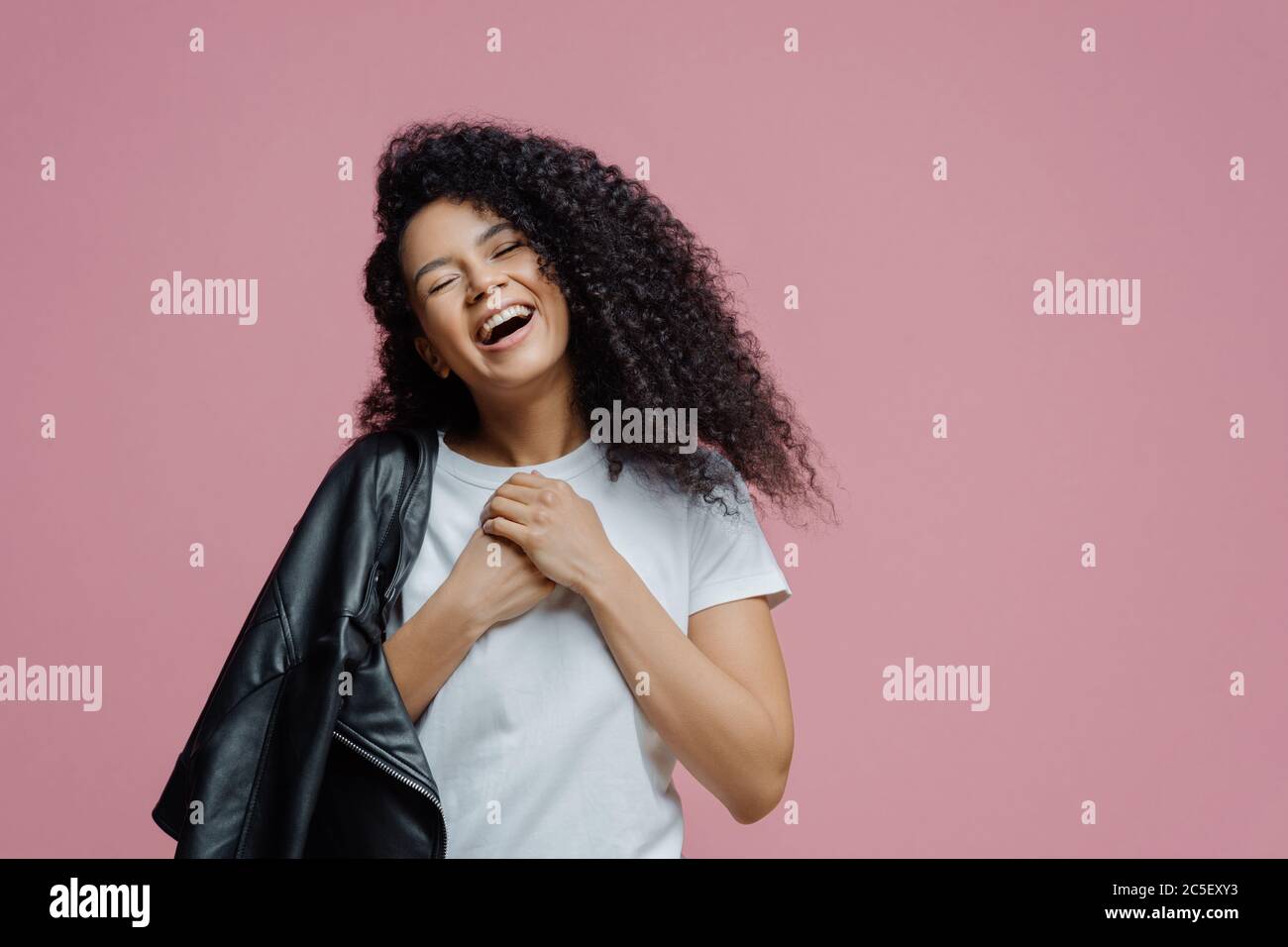 Une femme surjoyée à la peau foncée garde les mains ensemble et rit sincèrement, ferme les yeux et se sent très heureuse, montre des dents blanches parfaites, porte t t shirt et Banque D'Images