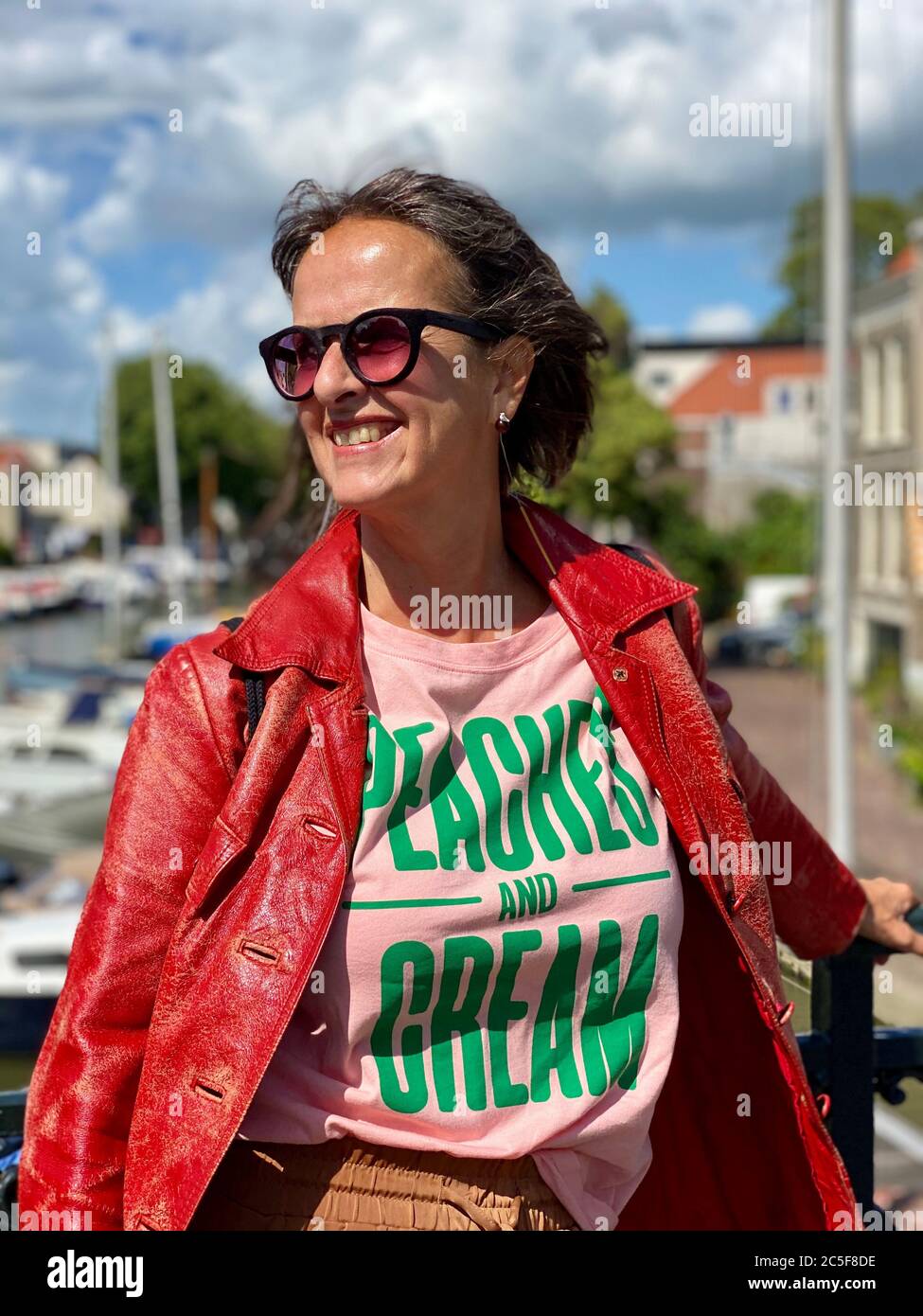 Portrait d'une femme mature et plus jeune, portant des lunettes de soleil et une veste en cuir rouge vintage, adossée à une main courante, montrant l'émotion souriante. Banque D'Images