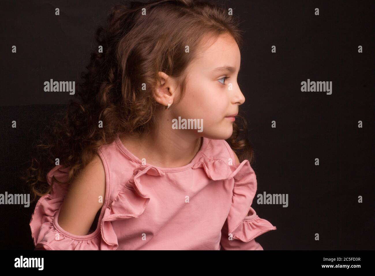 Portrait studio d'une petite fille aux cheveux longs et bouclés, dans une jupe rose et un short noir, assis sur une chaise de bureau, en soutenant son visage ses mains Banque D'Images