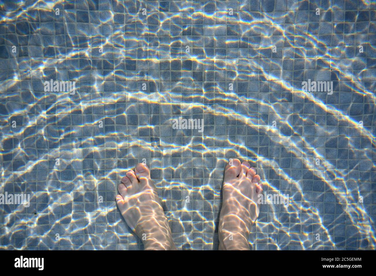 Les pieds blancs d'homme dans la piscine, le sol en mosaïque noir et gris clair mêlé les vagues sur la surface de l'eau sautent dans le soleil doré. Banque D'Images