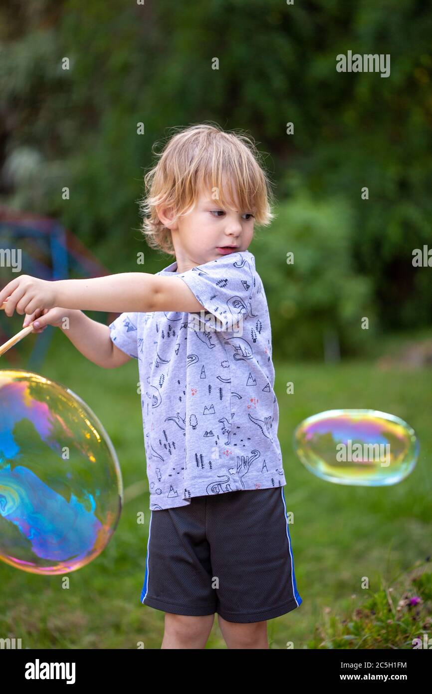 Jeune garçon d'âge préscolaire soufflant des bulles arc-en-ciel géantes dans un cadre de cour Banque D'Images