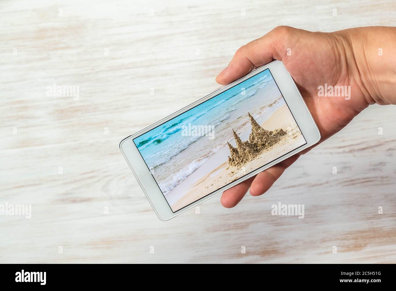 Téléphone portable avec photo de plage sur fond de bois. Smartphone à portée de main avec vue sur le sable et la mer. Main avec téléphone portable avec pho Banque D'Images