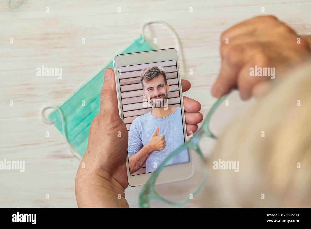 Femme avec cheveux blancs et lunettes regardant un gars dans le téléphone, masque médical sur la table. Un homme qui appelle sa mère par vidéo. Femme âgée effectuant un appel vidéo Banque D'Images