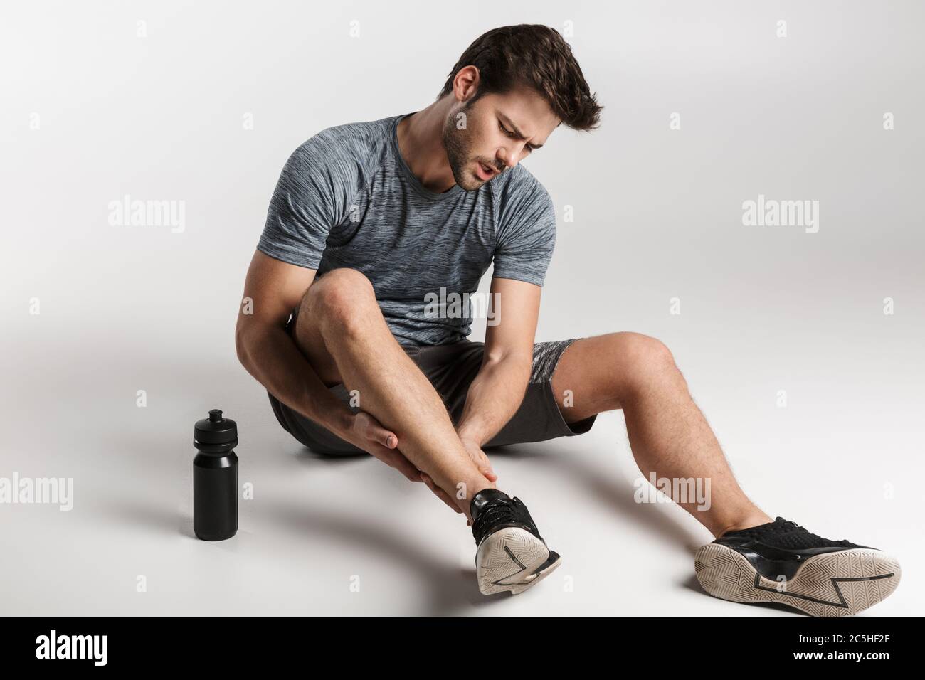 Jeune sportif fatigué reposant sur un sol isolé sur fond gris, souffrant de douleurs à la cheville Banque D'Images