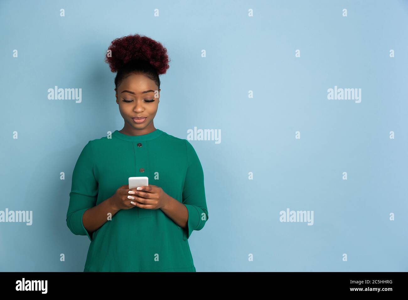 Téléphone intelligent à défilement. Portrait de la jeune femme afro-américaine isolé sur fond de studio bleu. Magnifique modèle féminin. Concept des émotions humaines, expression faciale, ventes, publicité. CopySpace. Banque D'Images