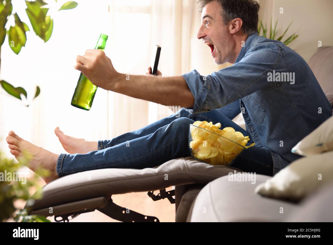 Homme regardant des sports à la télévision assis confortablement sur un canapé avec un en-cas avec bière et une télécommande dans ses mains. Banque D'Images