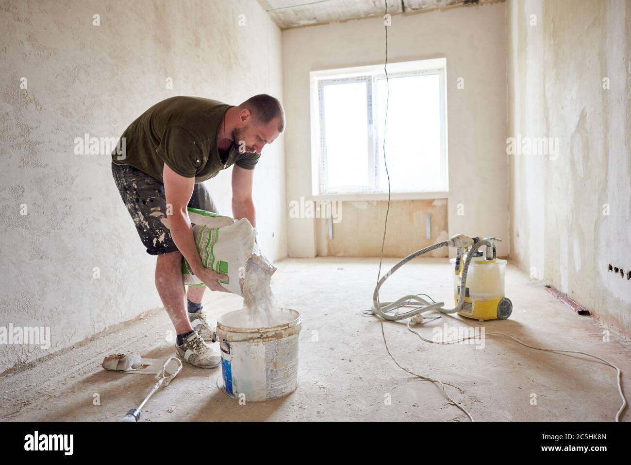 Le constructeur va mélanger le mastic en cours de réparation de la maison. Un homme dans une petite pièce préparé pour la réparation verse le mélange dans un seau à la lumière du jour contre une fenêtre Banque D'Images