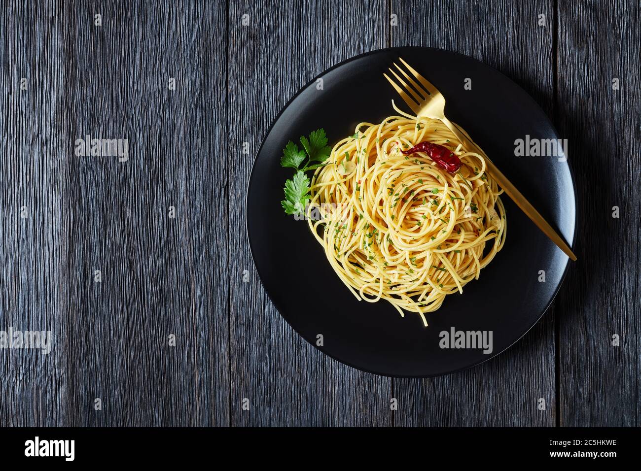 Spaghetti Alla colatura di alici, spaghetti à la sauce anchois, piment, ail et persil sur une assiette noire avec fourchette dorée sur un bois sombre Banque D'Images