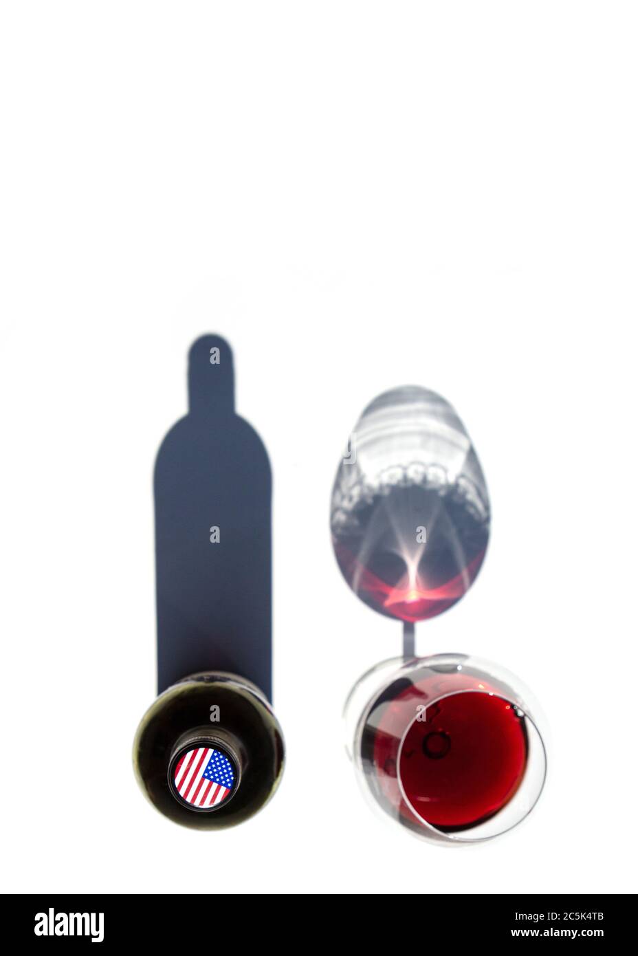 Une bouteille de vin et un verre de vin rouge jettent des ombres dures sur une table blanche. Le drapeau des États-Unis est représenté sur le bouchon de la bouteille. Banque D'Images