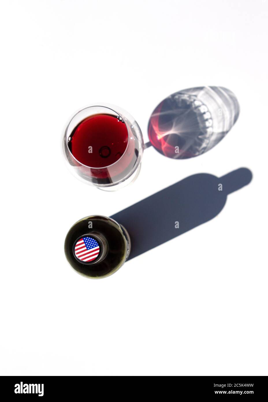 Une bouteille de vin et un verre de vin rouge jettent des ombres dures sur une table blanche. Le drapeau des États-Unis est représenté sur le bouchon de la bouteille. Banque D'Images