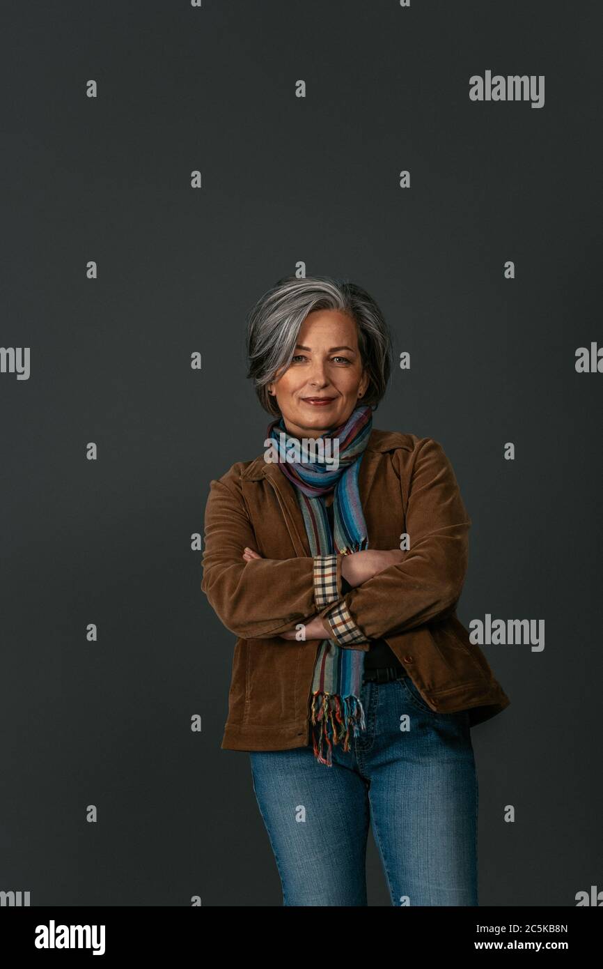 Belle femme créative et mûre sourit debout bras croisés sur le dos gris. Jolie femme blonde ou argentée dans un portrait décontracté. Image teintée Banque D'Images