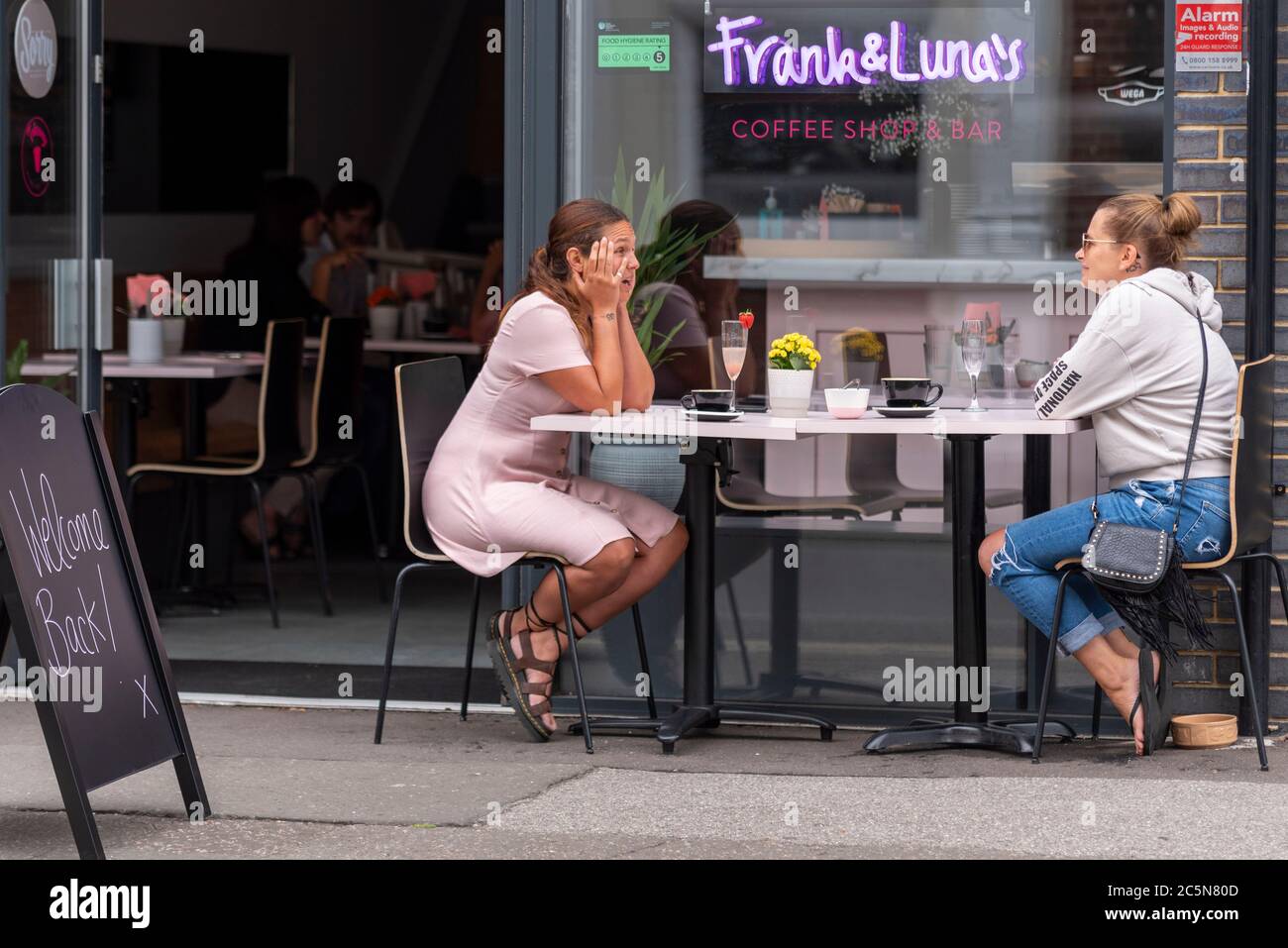 Southend on Sea, Essex, Royaume-Uni. 4 juillet 2020. Les pubs, restaurants et autres entreprises en Angleterre sont libres d'ouvrir leurs portes aux clients pour la première fois depuis le verrouillage du coronavirus COVID-19. Le gouvernement leur permet d'ouvrir à partir de 6h du matin comme facilité de verrouillage des mesures. Deux femmes blanches de race blanche dînant à l'extérieur du café restaurant avec panneau de bienvenue. Les gens mangent sur une table de pavement Banque D'Images