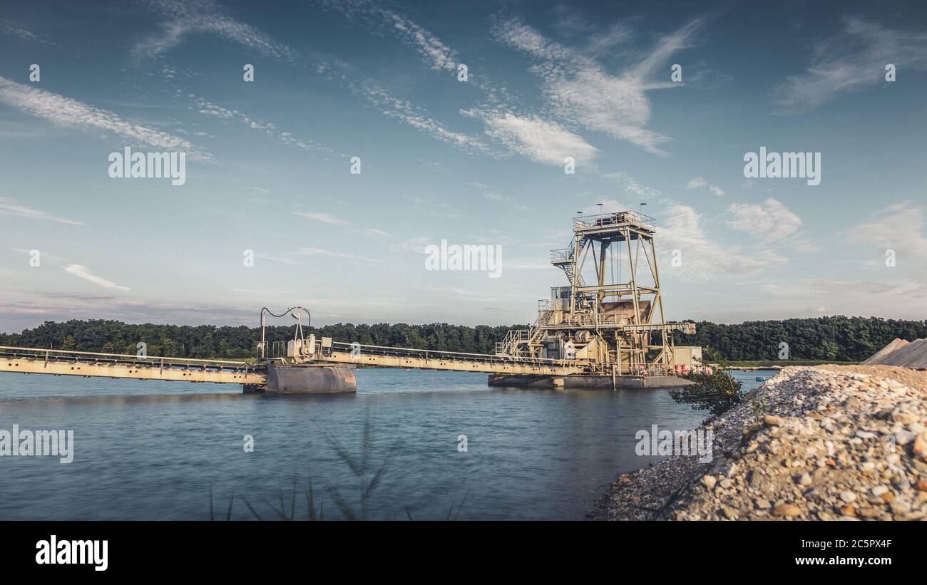 Tapis convoyeur industriel d'une installation d'extraction de gravier sur l'eau avec ciel sombre moody. Banque D'Images