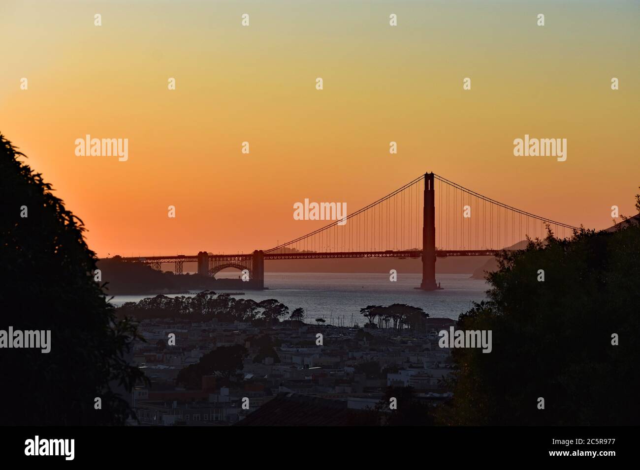 Coucher de soleil sur l'océan Pacifique avec le Golden Gate Bridge en silhouette avec ciel jaune et orange. Bâtiments et arbres au premier plan. San Francisco. Banque D'Images