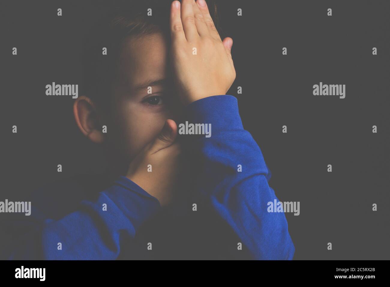 portrait d'un garçon aux cheveux sombres couvrant son visage avec ses mains sur un fond sombre Banque D'Images