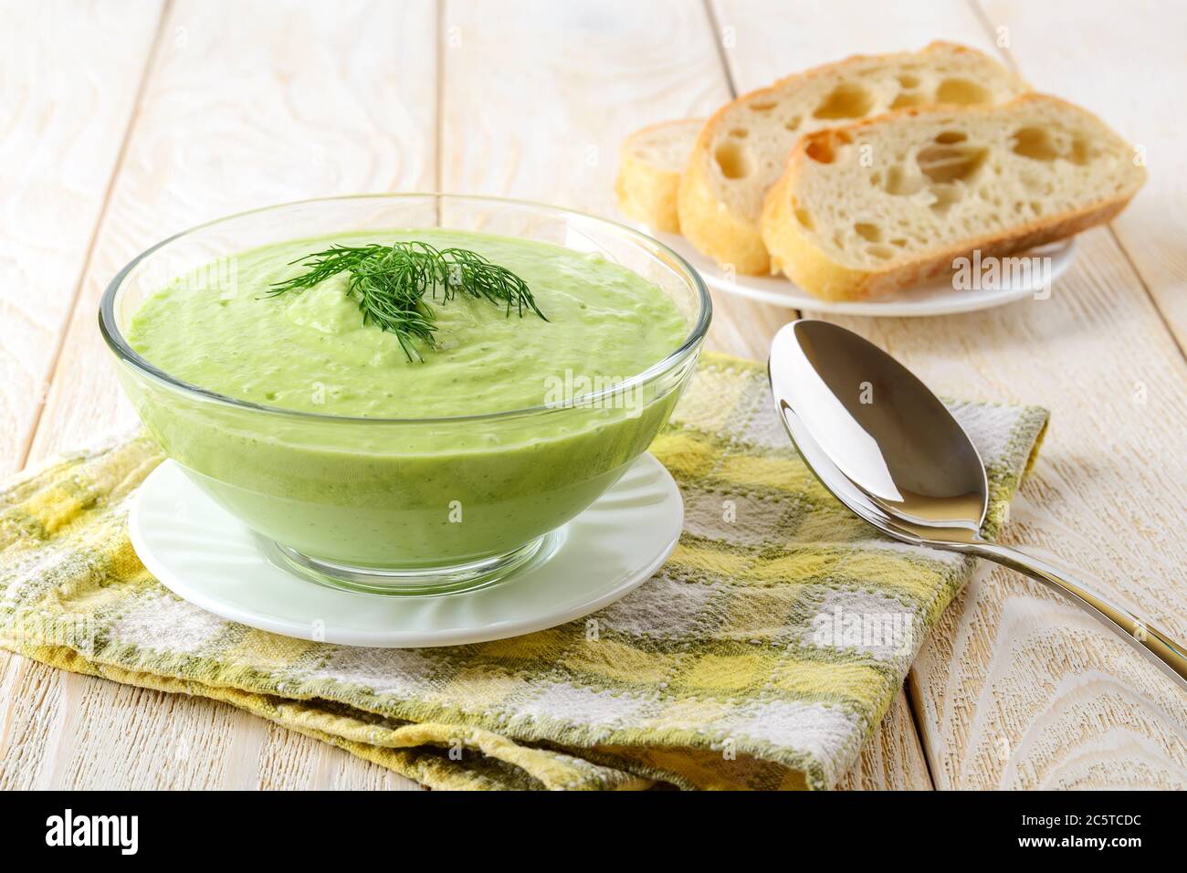 Gros plan de la purée de concombre froid dans un bol en verre, une cuillère et du pain sur une table en bois. Soupe de nourriture crue de yaourt grec, concombre, avocat, poivre Banque D'Images