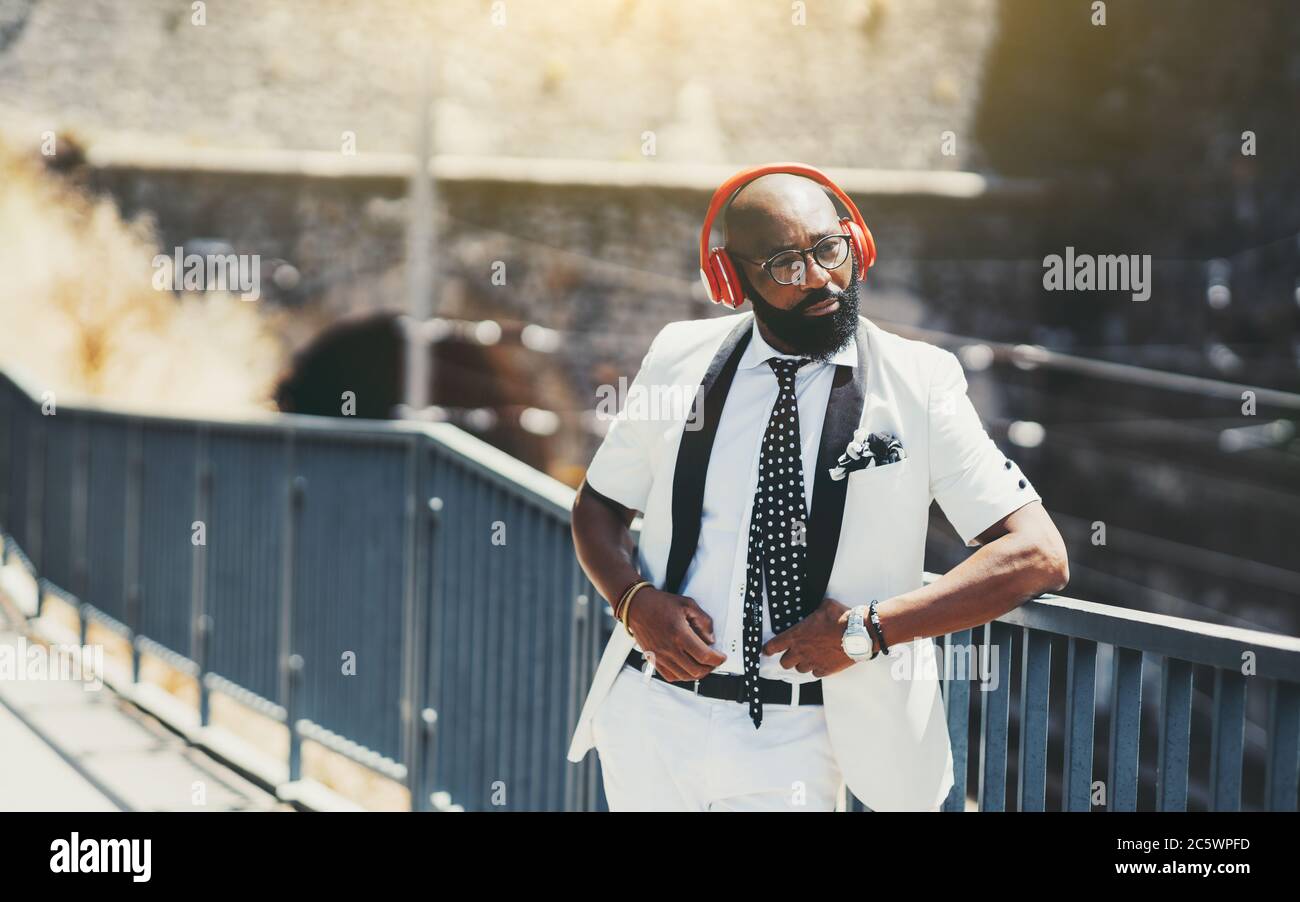 Un homme noir élégant à barbe chauve pour adultes, dans un costume blanc  chic avec cravate et casque sans fil rouge, écoute de la musique dans la  rue Photo Stock - Alamy