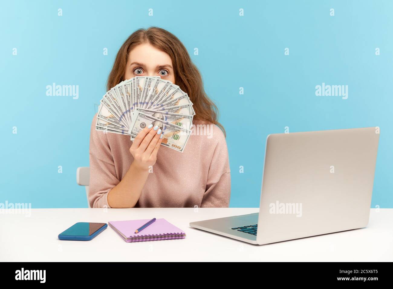 Drôle surprise femme employée assis sur le lieu de travail avec un ordinateur portable et de regarder des billets de dollars, avec un grand oeil émerveillé, choqué par le haut du sala Banque D'Images