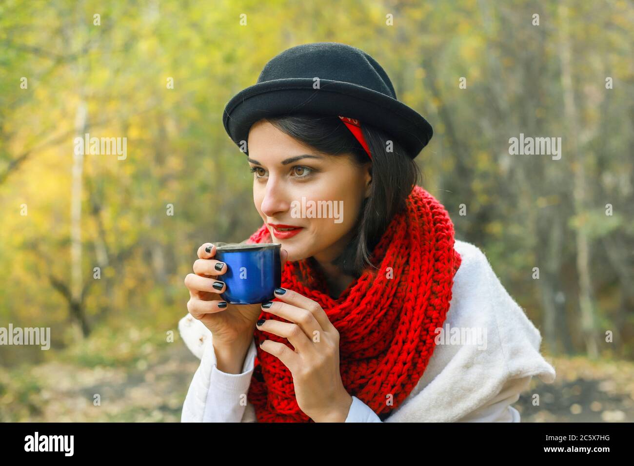 Belle jeune femme en manteau blanc, foulard rouge crochet et chapeau noir tient une tasse dans ses mains. Fête du thé dans la nature Banque D'Images