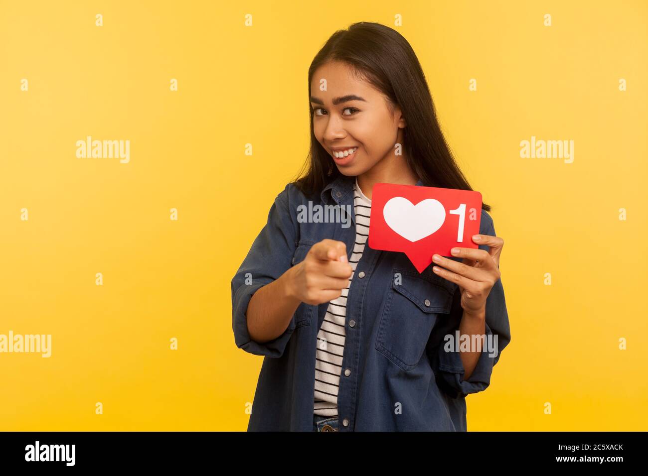 Hé, vous ! Portrait d'une fille ludique dans une chemise en denim pointant vers l'appareil photo et montrant le cœur comme une icône, symbole de notification de suiveur, cliquez sur le bouton pour social Banque D'Images