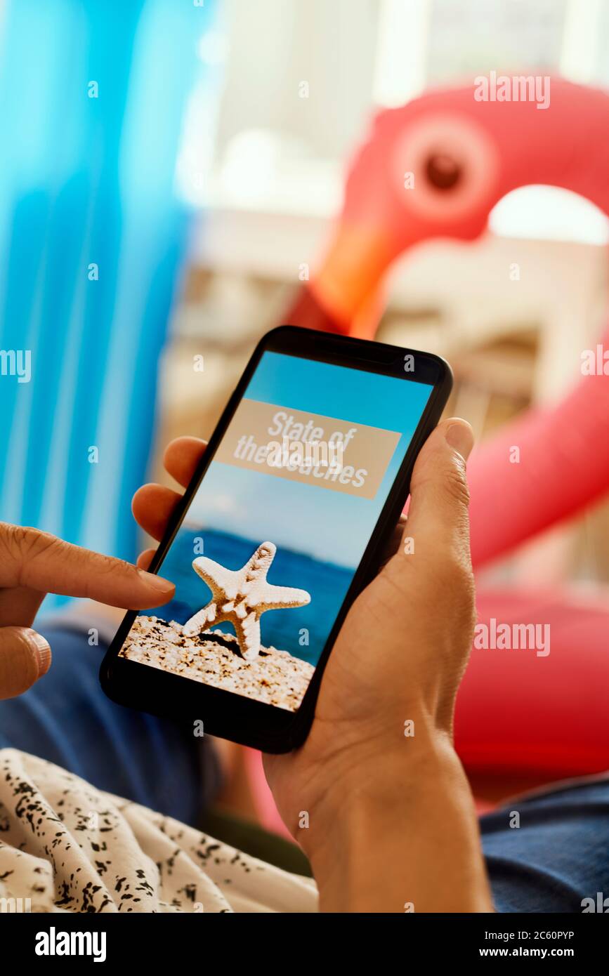 jeune homme, portant des vêtements d'été décontractés, assis sur un fauteuil, ayant son smartphone entre ses mains avec le texte état des plages dans son écran, Banque D'Images