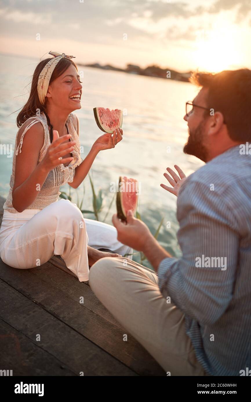 jeune couple assis près de l'eau parlant, souriant, riant, mangeant de pastèque Banque D'Images