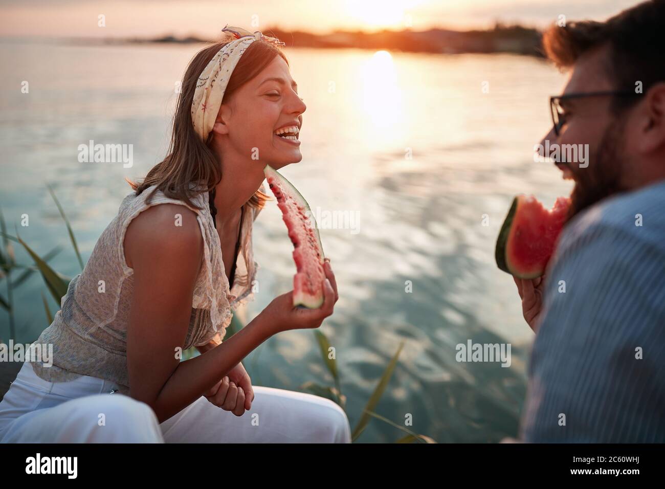 jeune couple mangeant de la pastèque, assis près de l'eau, souriant, riant, parlant Banque D'Images
