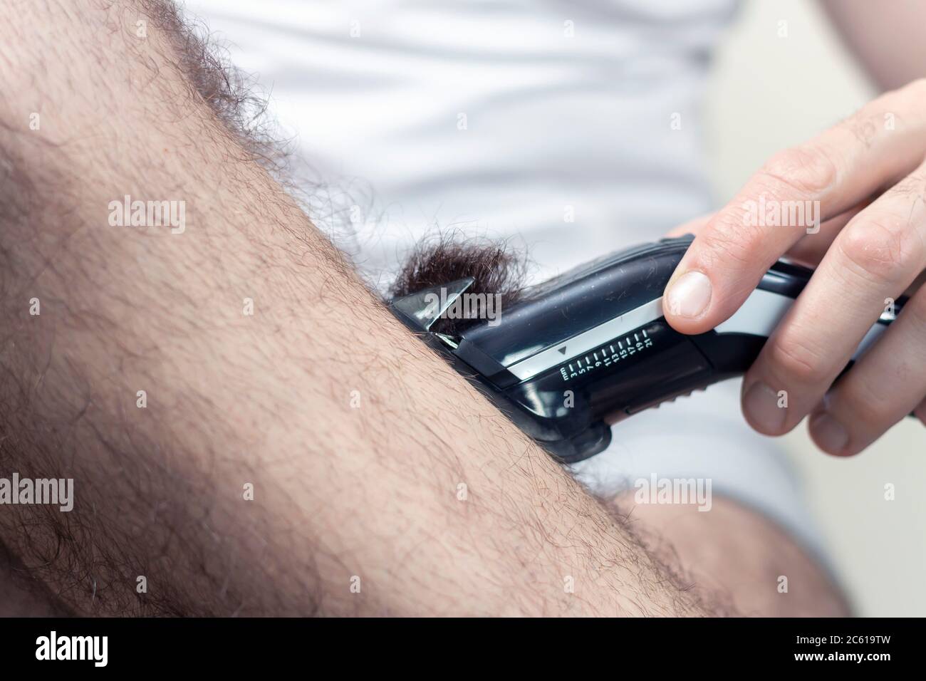 La main de l'homme tient une tondeuse à cheveux électrique et coupe de longs cheveux sombres sur ses jambes. Banque D'Images