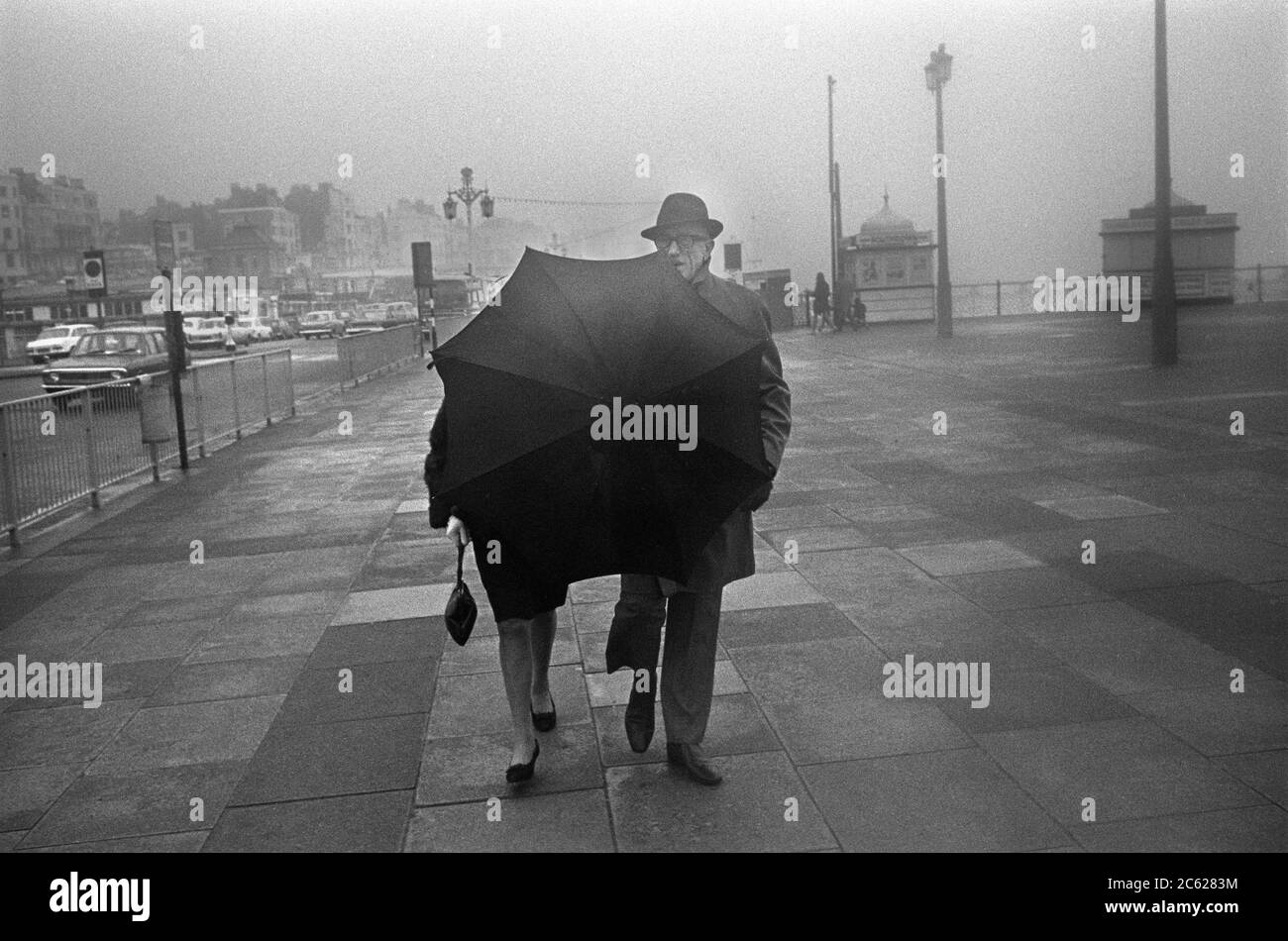 Parasol Brighton front de mer promenade Sussex Angleterre 1960s. Couple d'âge moyen marchant pendant le mauvais temps traditionnel anglais de la mer 1969. ROYAUME-UNI HOMER SYKES Banque D'Images