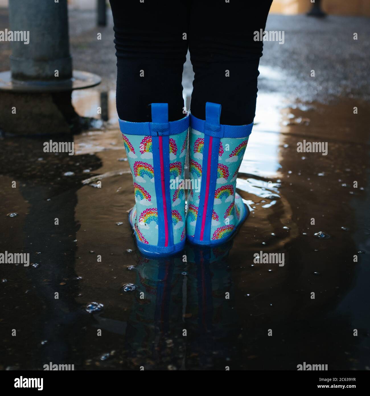 Fille jouant sur une scène urbaine sautant au-dessus de flaques d'eau avec des bottes bleu arc-en-ciel Banque D'Images