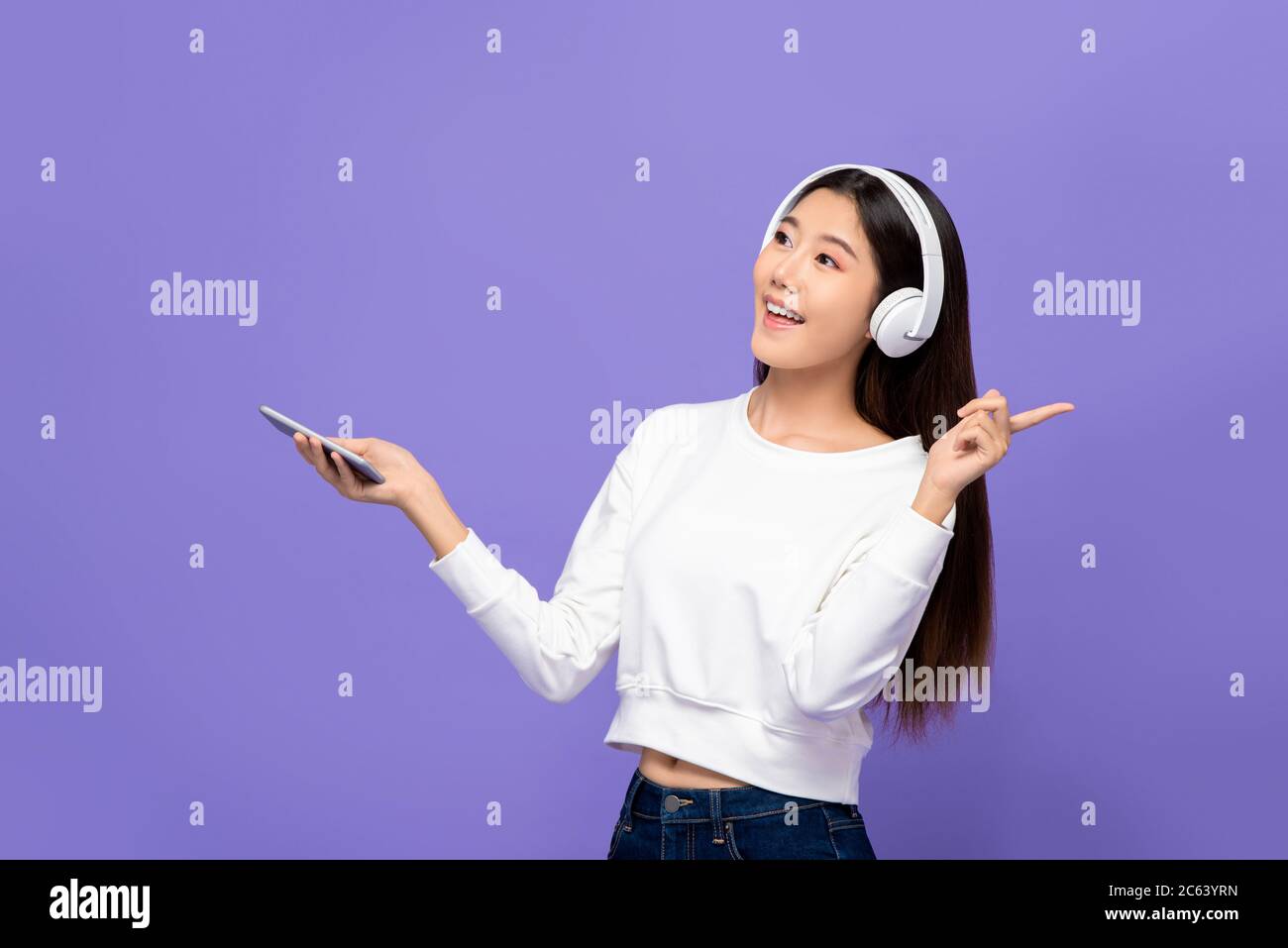Portrait d'une jeune femme asiatique souriante écoutant de la musique sur un téléphone mobile à l'aide d'un casque sans fil dans un fond de studio violet isolé Banque D'Images