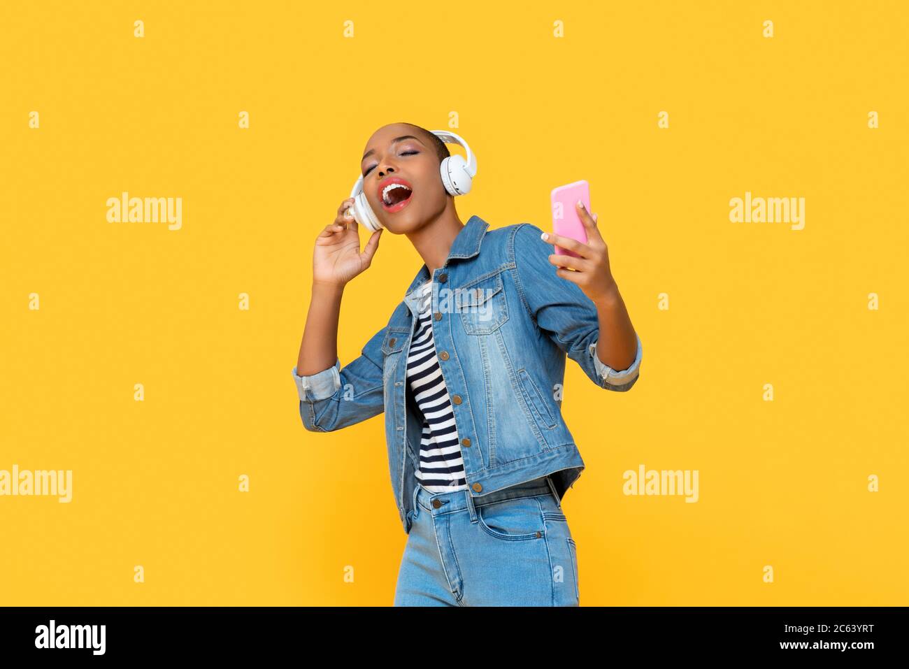 Bonne femme afro-américaine portant un casque à l'écoute de musique en ligne dansant et chantant sur fond jaune isolé Banque D'Images