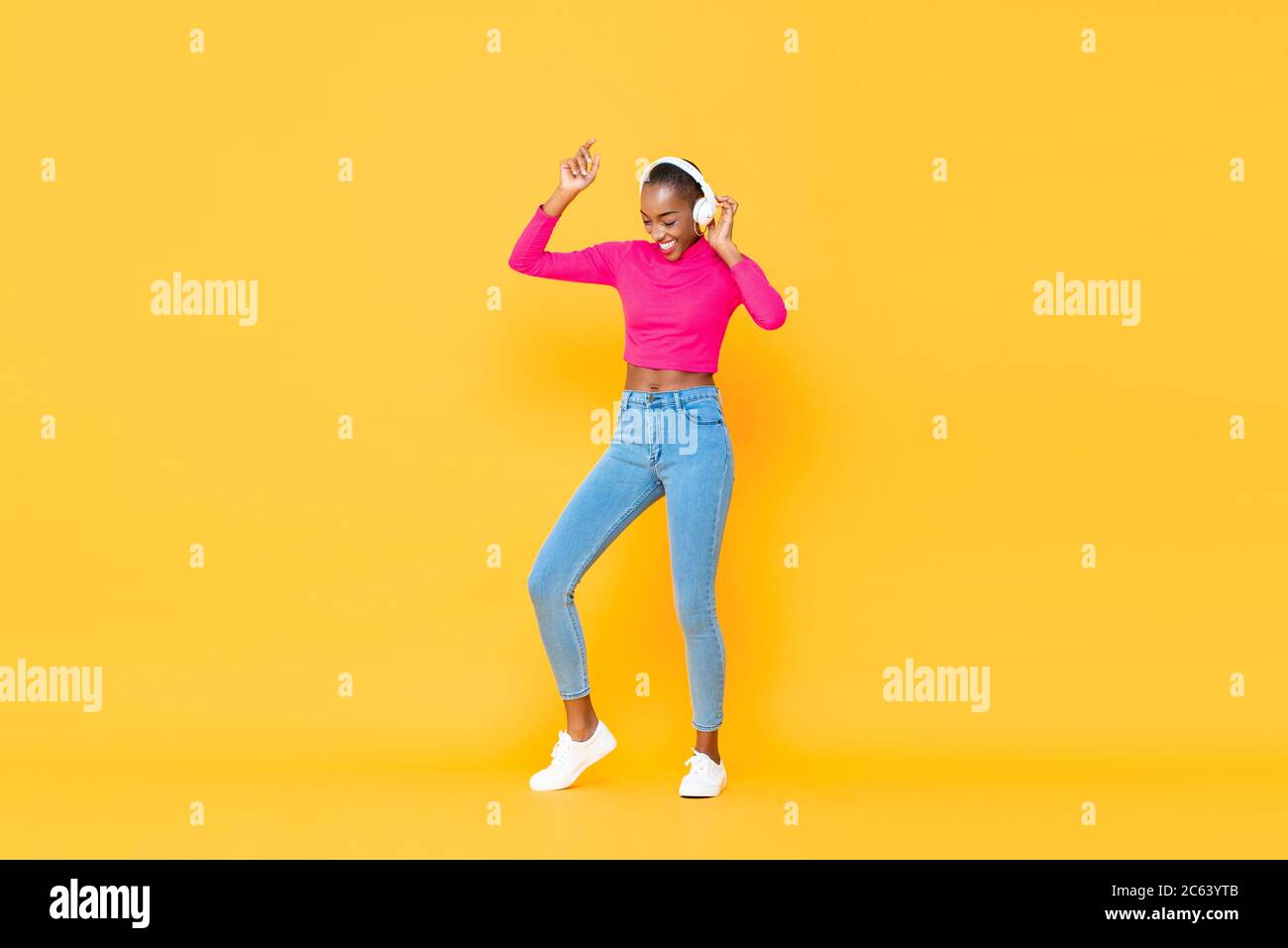 Bonne femme afro-américaine portant un casque, écoutant de la musique et dansant sur fond jaune coloré isolé Banque D'Images