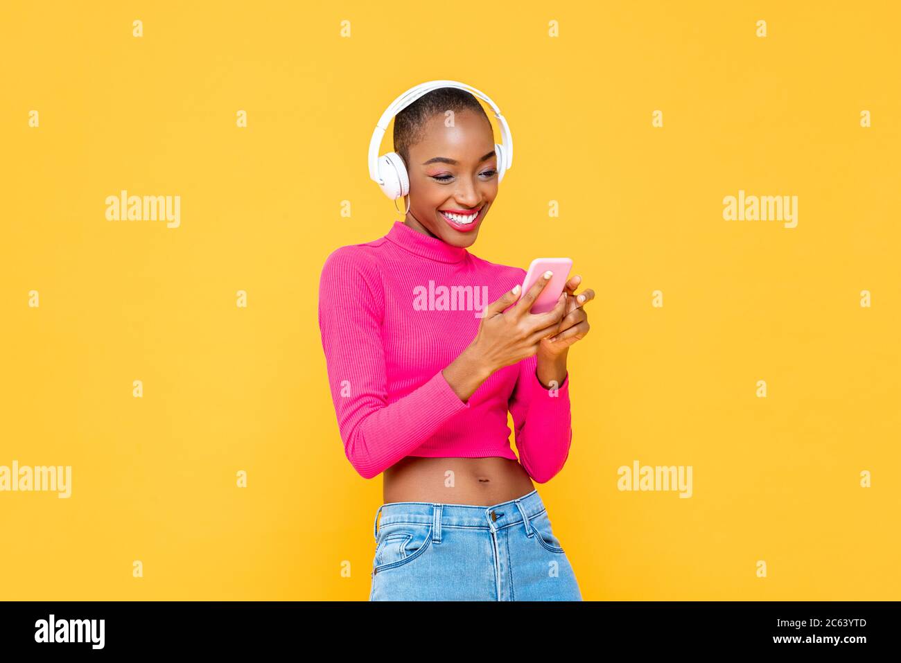 Femme afro-américaine heureuse et tendance, avec casque, qui écoute de la musique depuis un smartphone sur fond de studio isolé jaune coloré Banque D'Images
