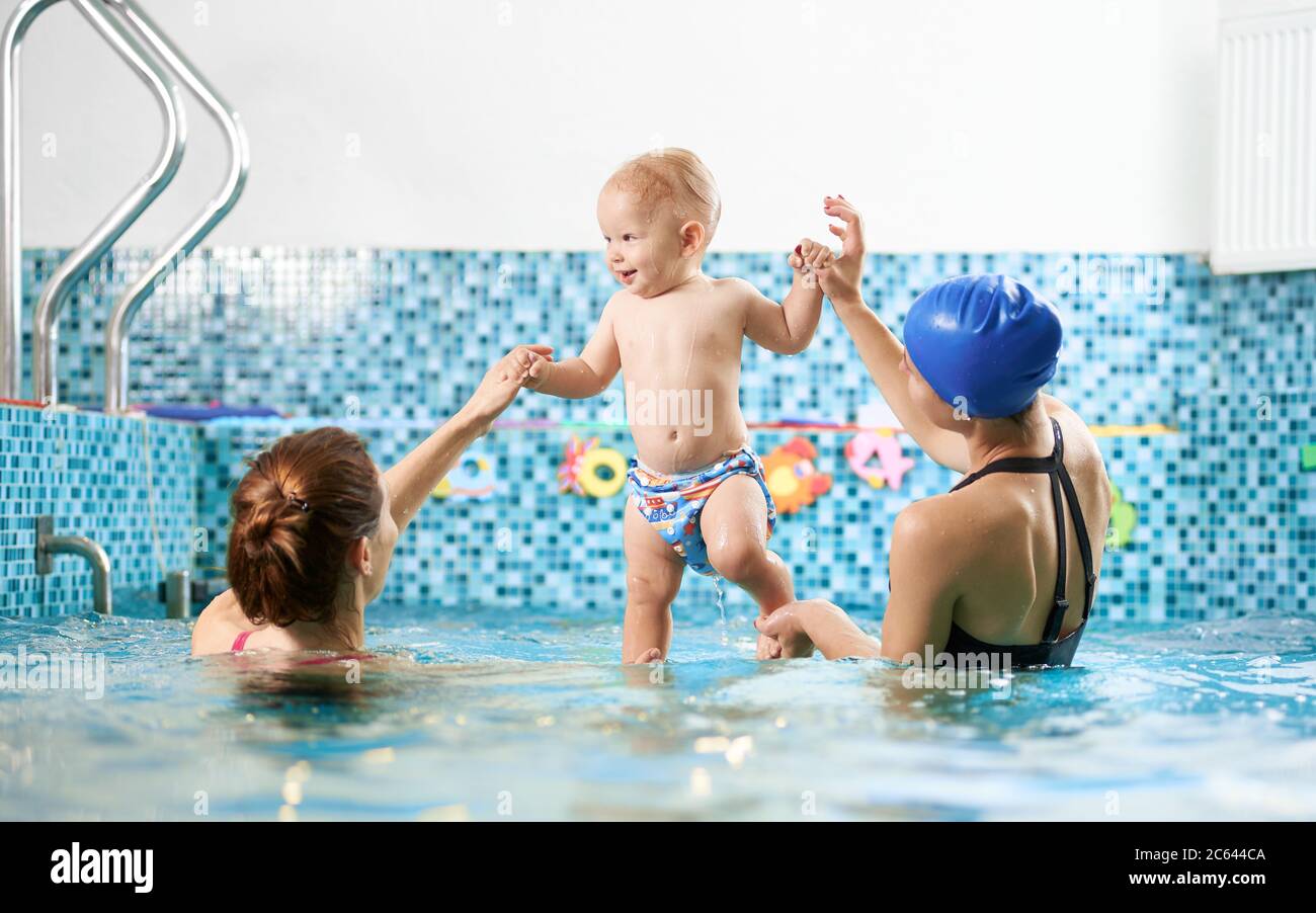 Vue Arriere De Deux Femmes Dans La Piscine Aidant Un Petit Garcon A Garder L Equilibre Dans L Eau Bebe Souriant Est Debout Dans Leurs Mains Concept De Sport Pour Enfants Photo Stock