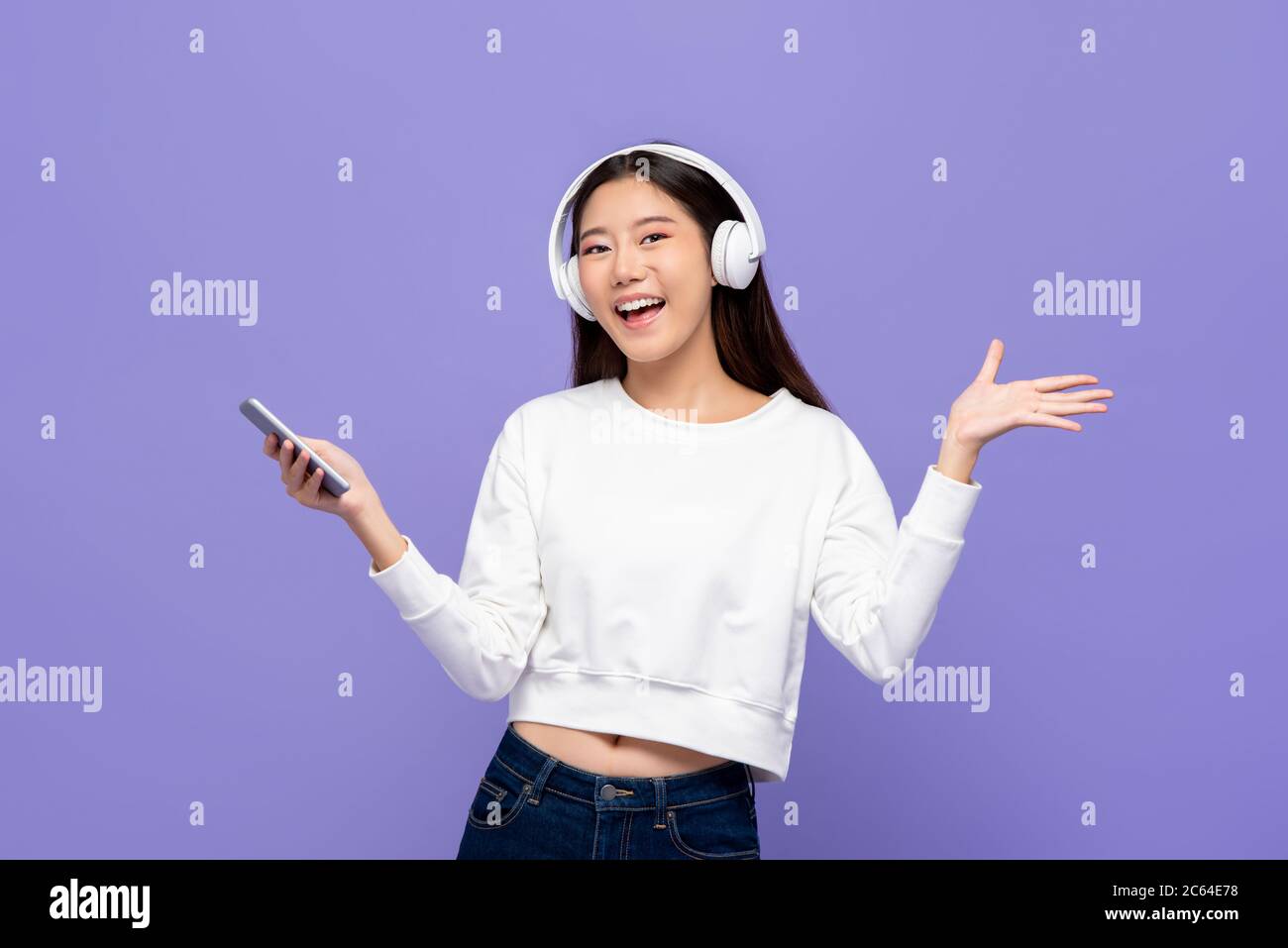 Jeune femme asiatique portant un casque d'écoute, écoutant de la musique depuis un smartphone et ouvrant la main isolée sur fond violet clair Banque D'Images