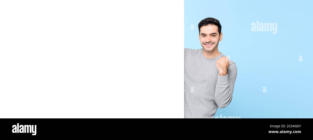 Souriant beau homme caucasien levant poing derrière le tableau blanc vide sur fond de bannière bleu clair Banque D'Images