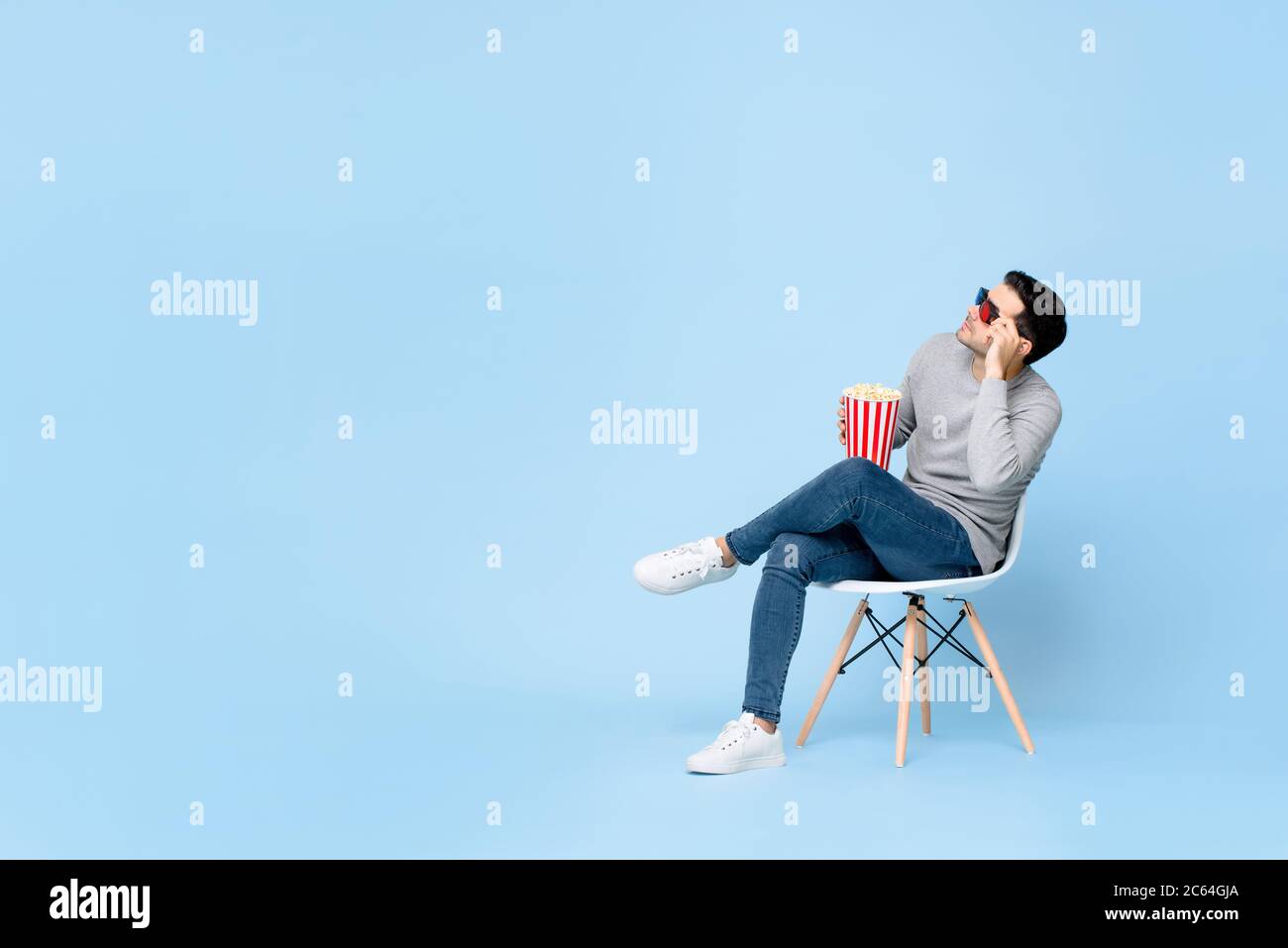 Jeune homme caucasien avec pop-corn et lunettes 3d regardant l'espace vide tout en étant assis sur une chaise isolée sur fond bleu clair Banque D'Images
