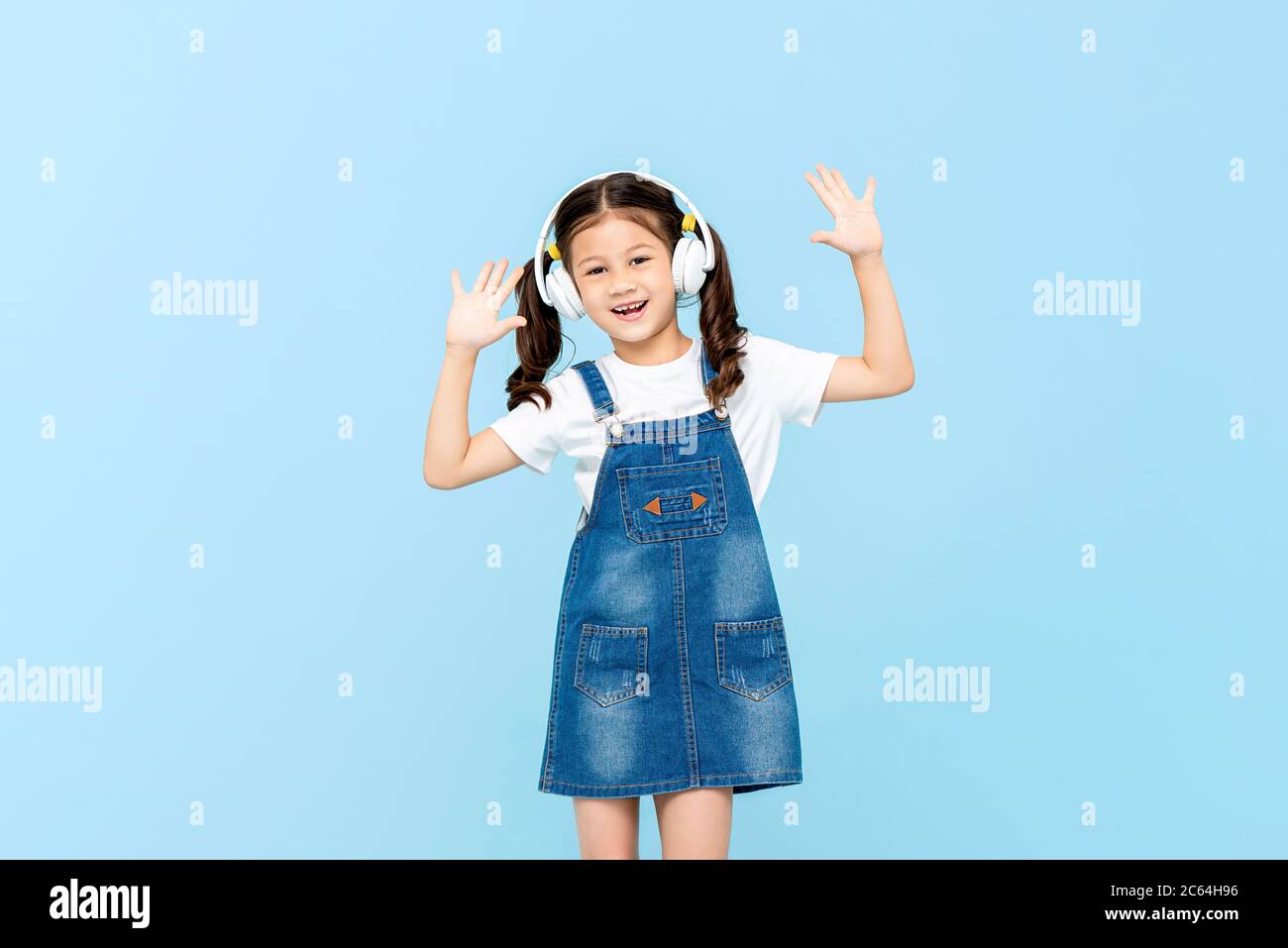Portrait amusant de la jeune fille asiatique heureuse écoutant de la musique dans les écouteurs faisant un geste de main levée dans le studio isolé bleu fond Banque D'Images