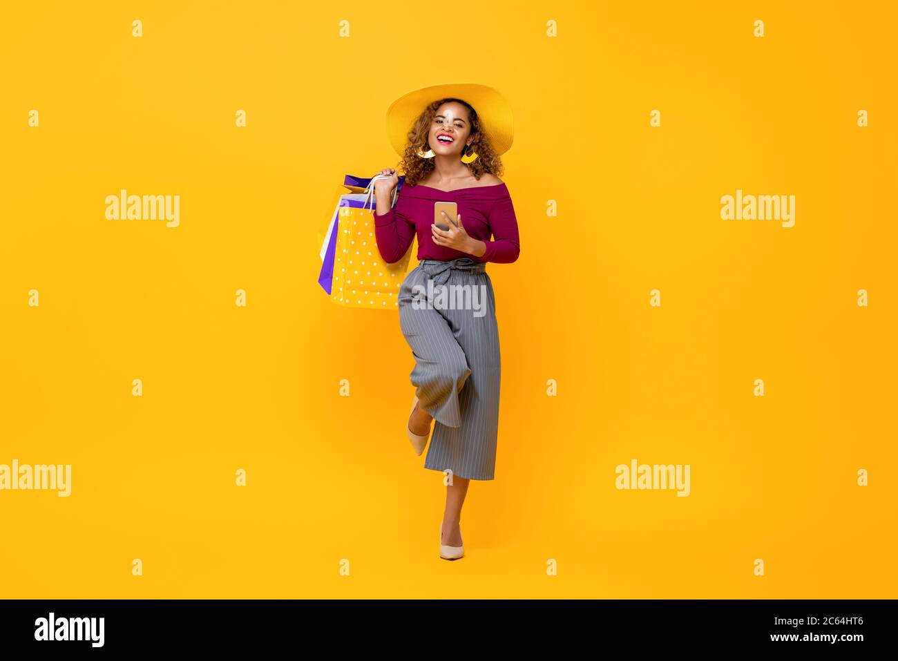 Portrait complet de jeune femme afro-américaine attrayante et heureuse tenant des sacs à provisions et un téléphone portable avec une jambe relevée dans le studio vous isolé Banque D'Images