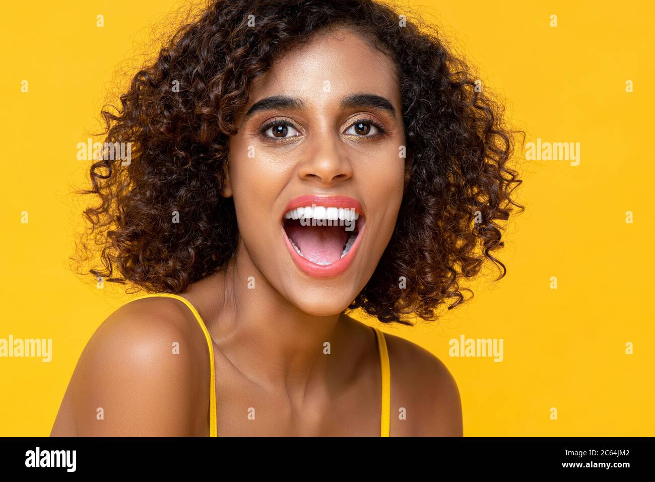 Gros plan portrait d'une femme afro-américaine heureuse regardant l'appareil photo avec la bouche ouverte dans un arrière-plan isolé de studio jaune Banque D'Images