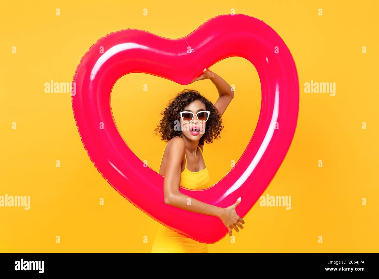 Portrait d'une femme afro-américaine surprise portant des lunettes de soleil tenant un ballon en forme de coeur sur fond jaune isolé de studio Banque D'Images