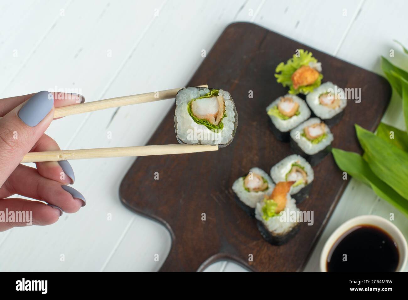 Femme tenant main avec un rouleau de sushi maki frais avec des baguettes en bois. Des petits pains à sushis sont servis sur un panneau en bois sombre. Fond gris avec feuilles vertes Banque D'Images
